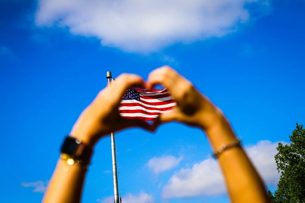 Someone making a heart shape with both hands and an American flag is waving in the background.