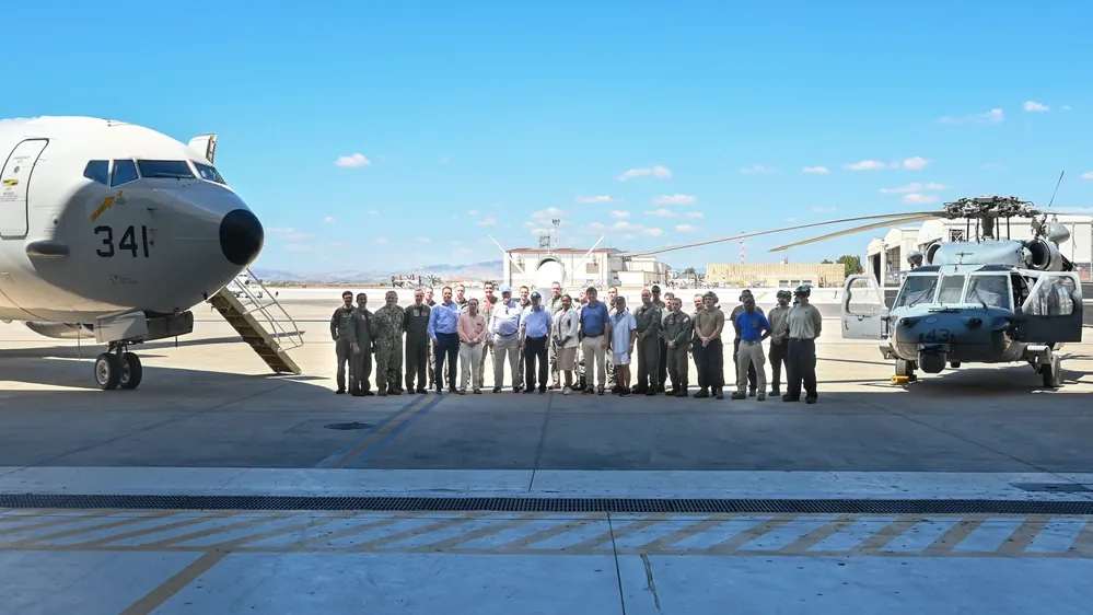 Group photo of U.S. military personnel and a congressional delegation standing on an airfield between a large Navy aircraft and a helicopter under clear skies.